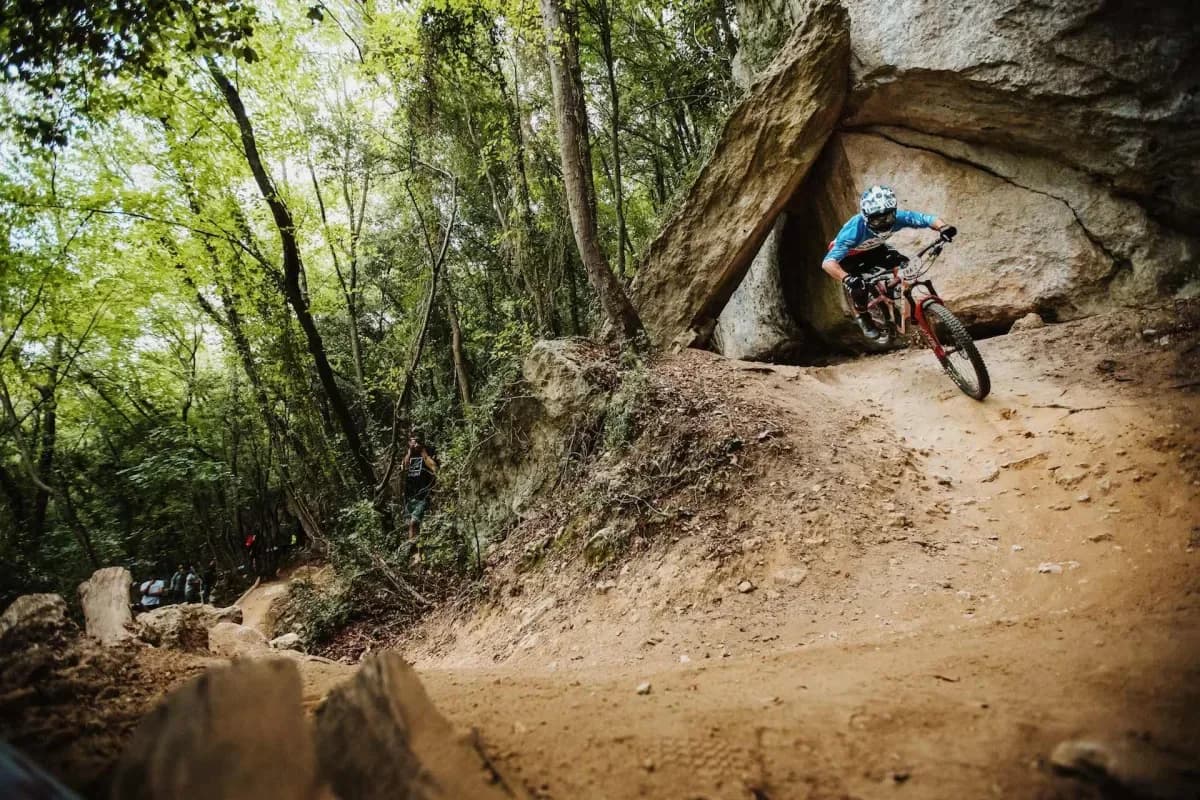 A rider drops past a limestone boulder on a dusty trail through the woods